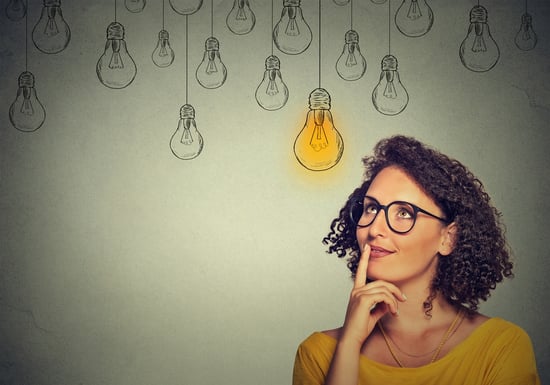 Thinking woman in glasses looking up with light idea bulb above head isolated on gray wall background.jpeg